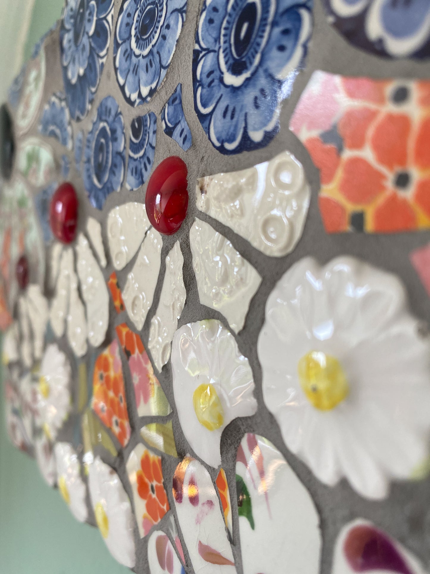 Close up of a small mosaic flower garden, with white; white and patterned, orange petals, and a blue sky with cut outs of crockery. Red and dark purple glass nuggets.