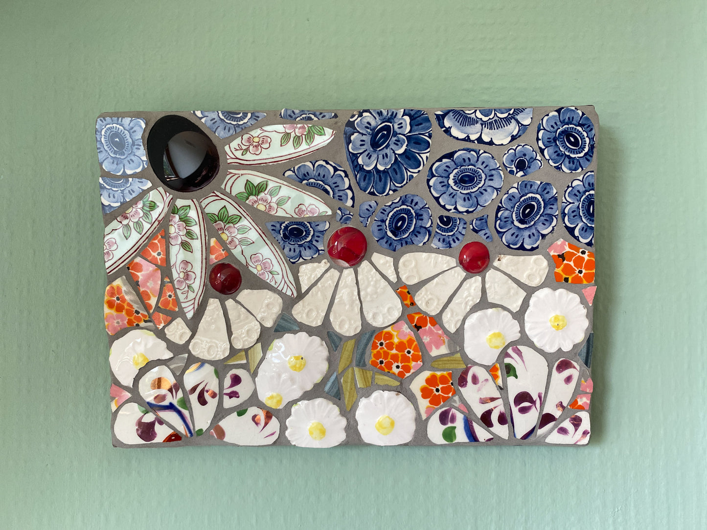 small mosaic flower garden, with white; white and patterned, orange petals, and a blue sky with cut outs of crockery. Red and dark purple glass nuggets. Shown against a pale green background