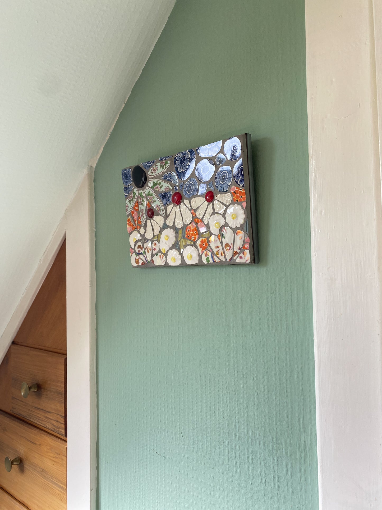 small mosaic flower garden, with white; white and patterned, orange petals, and a blue sky with cut outs of crockery. Red and dark purple glass nuggets. Shown on an internal wall