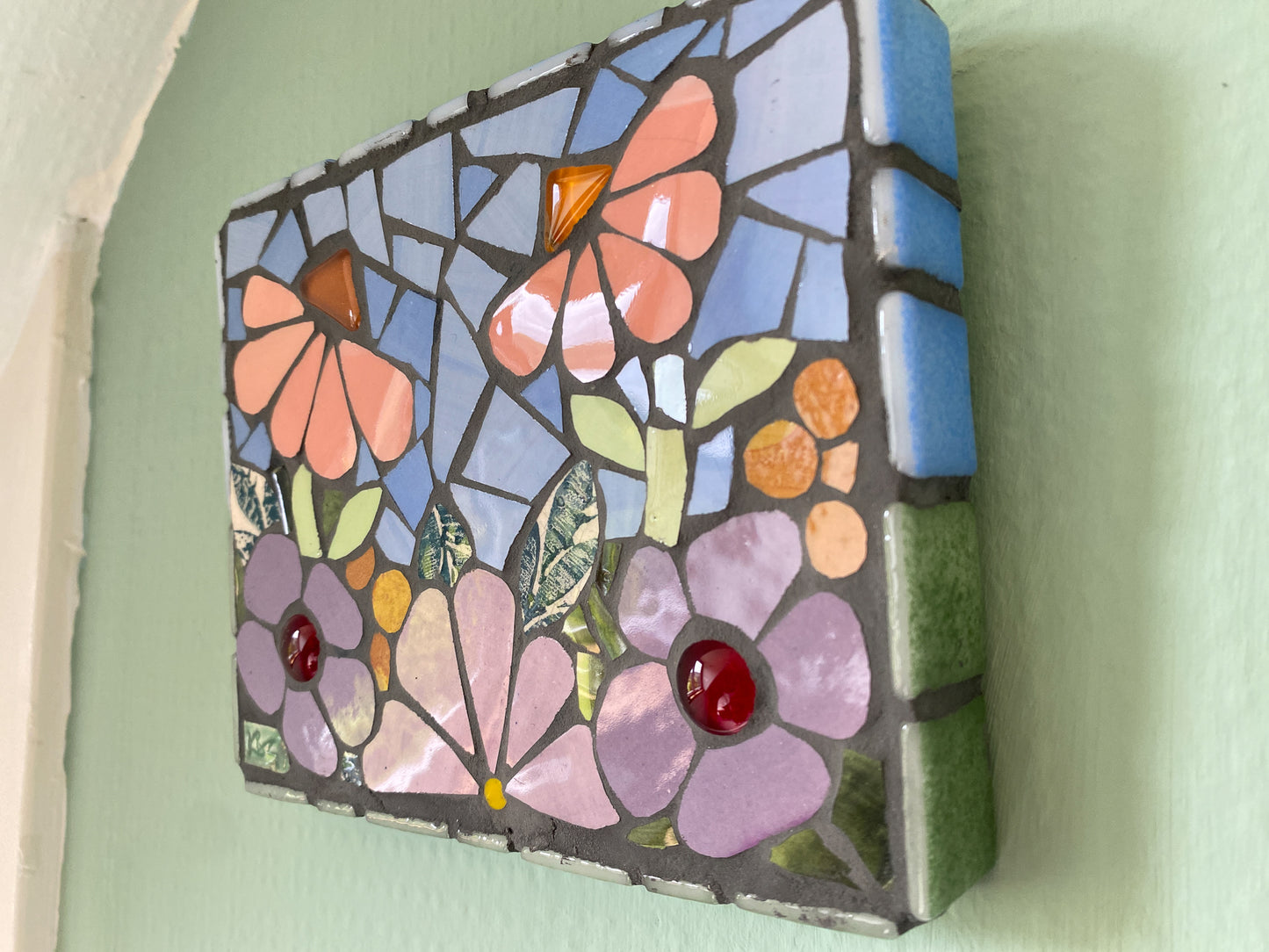 small flower garden mosaic with purple and peach toned flowers, patterned leaves blue sky, and orange and red glass nuggets. Shown on a green background At an angle, showing the tiled edging