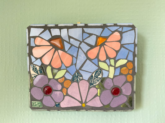 small flower garden mosaic with purple and peach toned flowers, patterned leaves blue sky, and orange and red glass nuggets. Shown on a green background