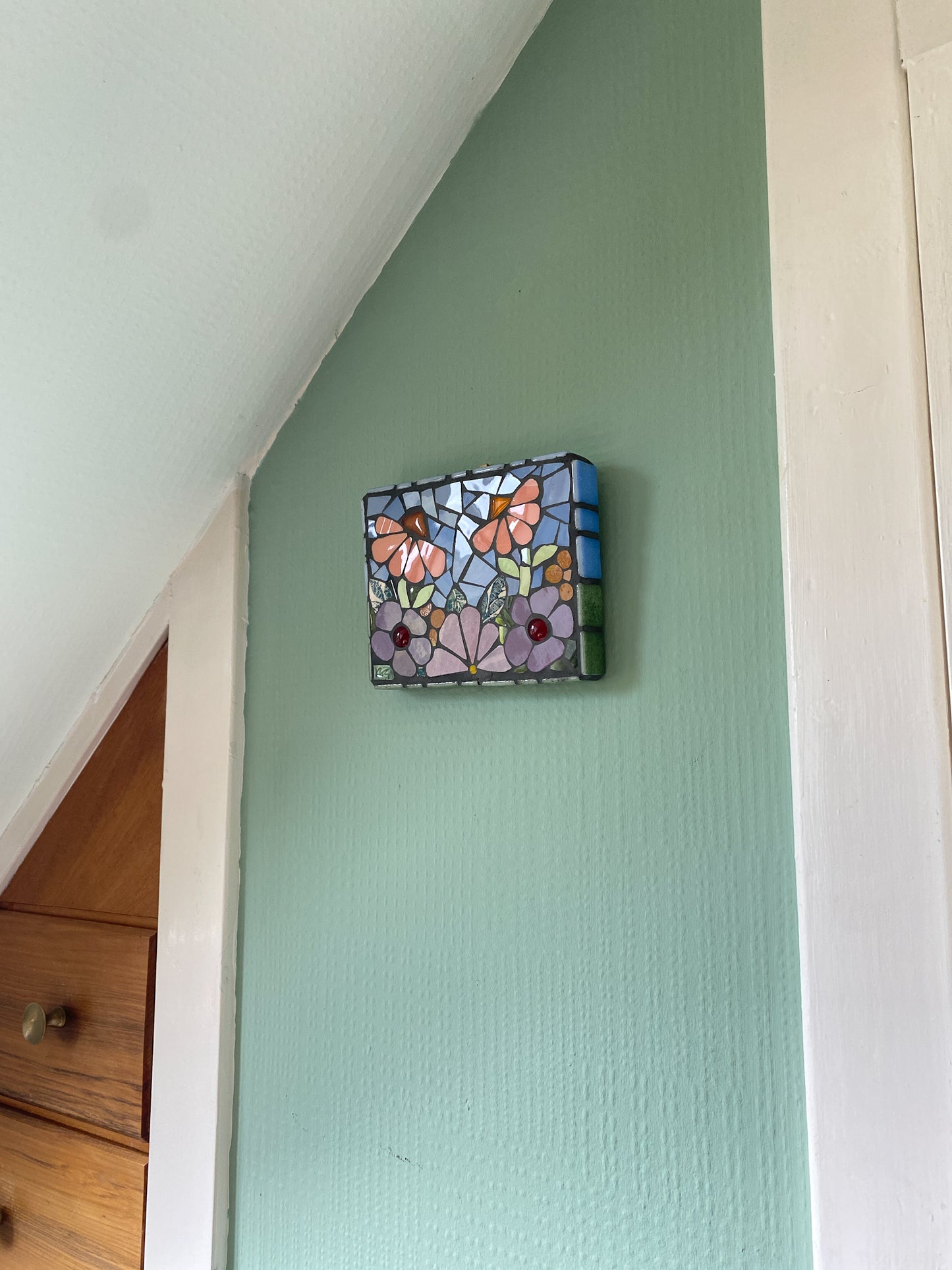 small flower garden mosaic with purple and peach toned flowers, patterned leaves blue sky, and orange and red glass nuggets. Shown on an internal green wallbackground