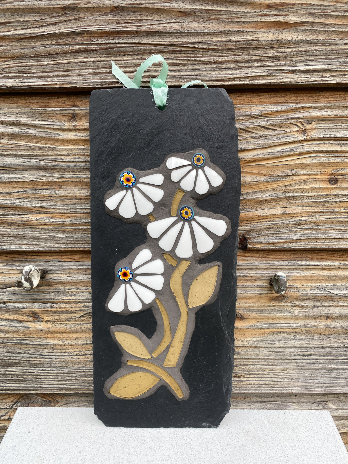 A rectangular black slate wall hanging ornament featuring a raised floral design with white petals, tan leaves and stems, and small multicolor flower centers. A mint green ribbon loop is attached at the top, and the ornament is photographed against a rustic wooden plank background.