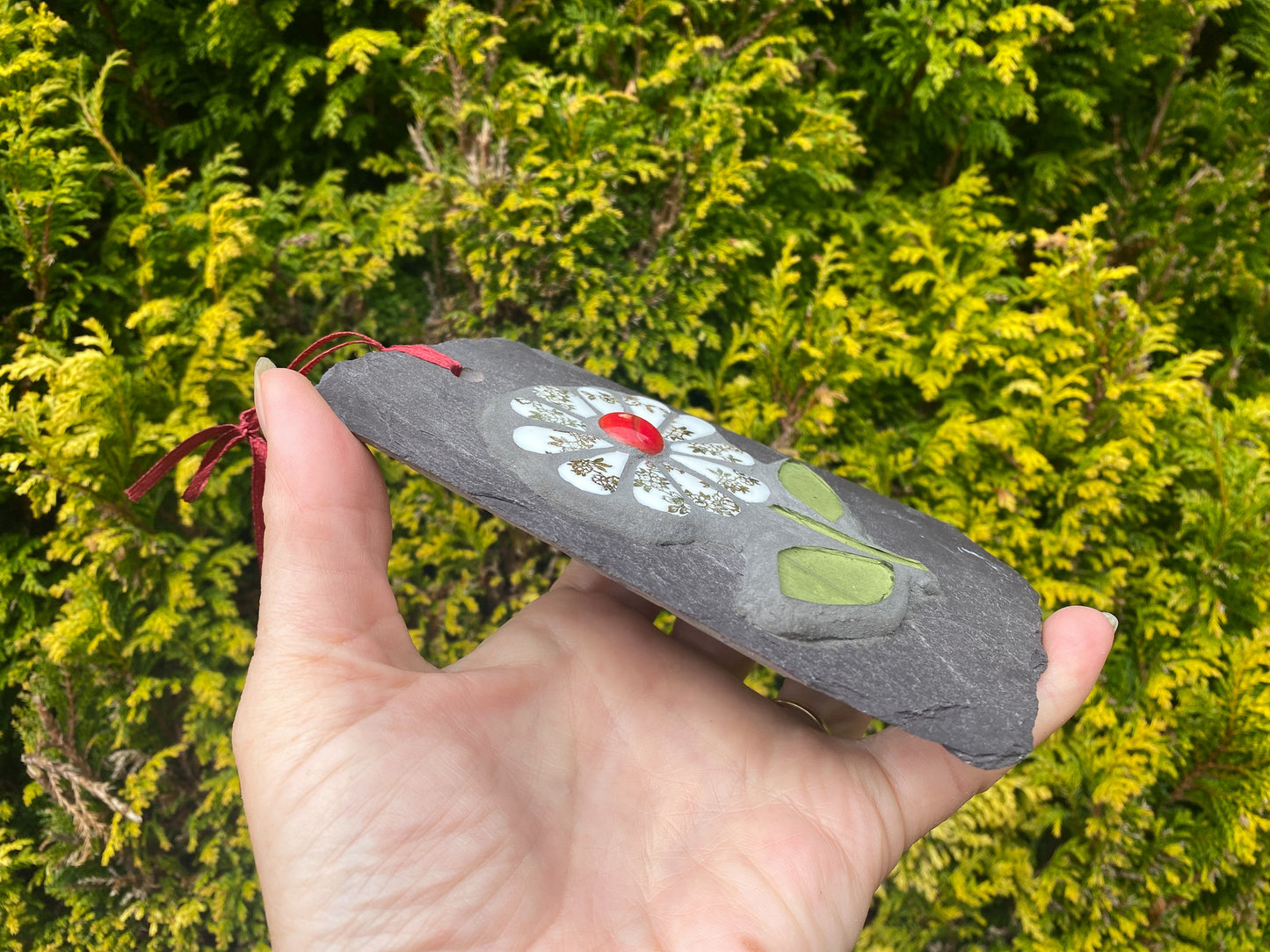gilt and white petalled daisy mosaic on slate,, showing the depth of the mosaic, hand held with an evergreen tree behind