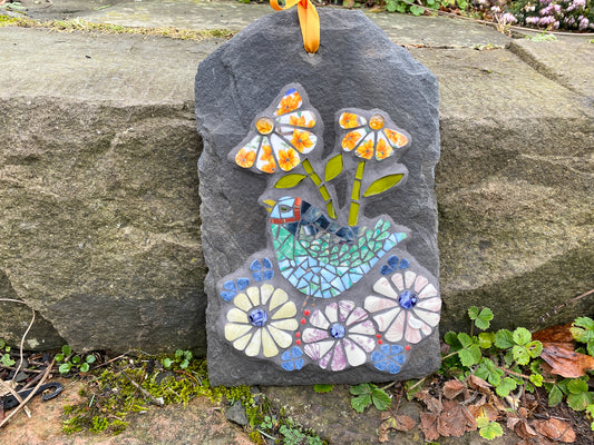 mosaic on slate with a green and blue bird, with purple and yellow flowers.  The petals are patterned from the crockery used.  Photographed on a stone step