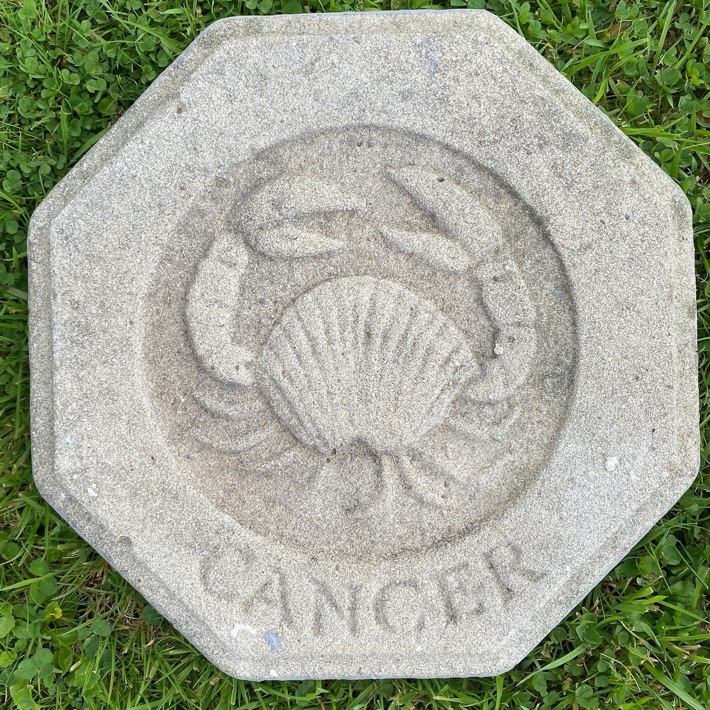 An octagonal gray stone or concrete garden marker featuring a carved crab design and the word 'CANCER' around the top edge, placed on green grass.