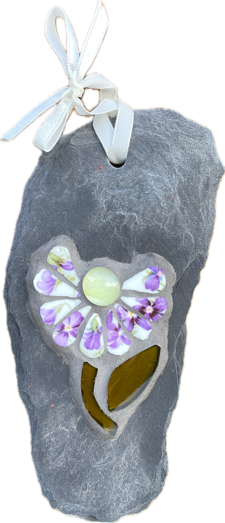 small daisy in mosaic with violet flowers on the petals. a pale yellow glass nugget at the centre of the flower, dark green stem and leaf. on a pale grey thin slate, shown on a white background