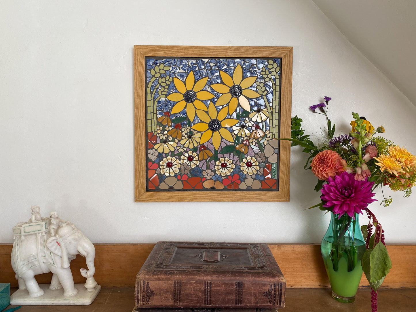 Framed mosaic artwork of flowers on a wall with a decorative vase and elephant statue below.