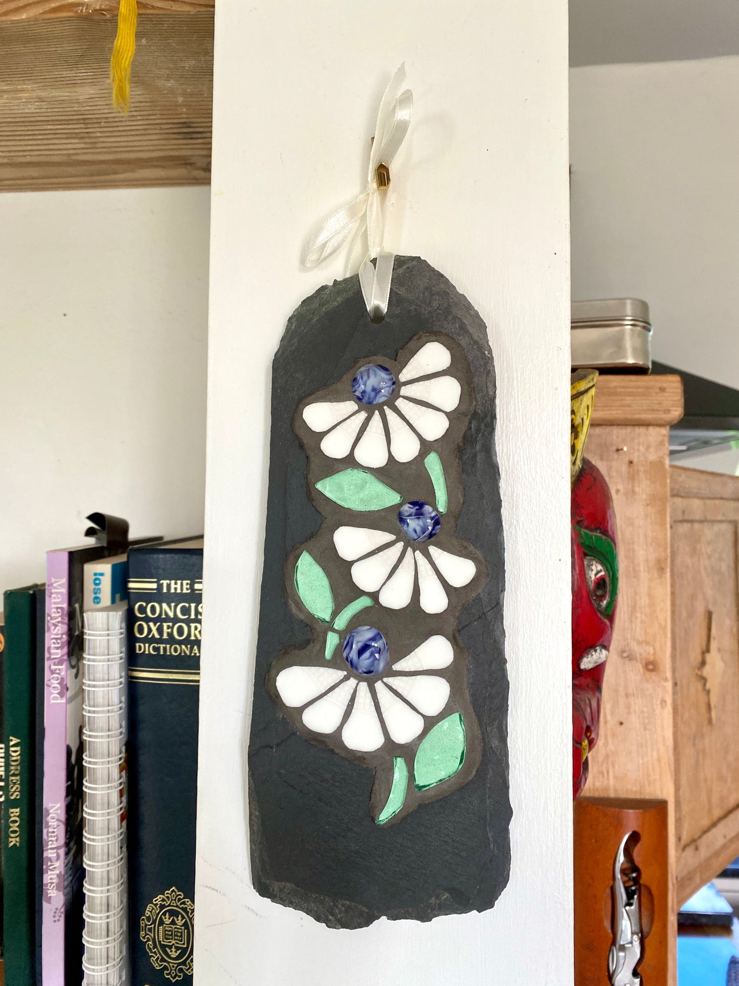 Decorative slate with floral design in mosaic hanging on a white wall next to books.