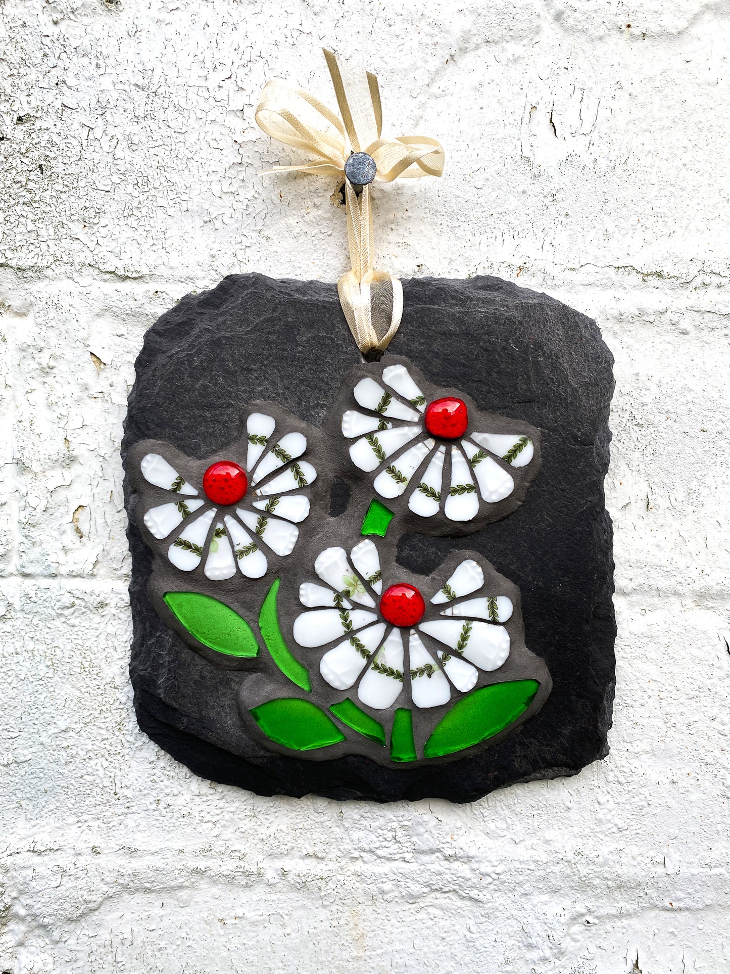 three white daisy flowers, with a green leafy detail, red glass nuggets and bright green leaves in mosaic on slate against a distressed white brick background