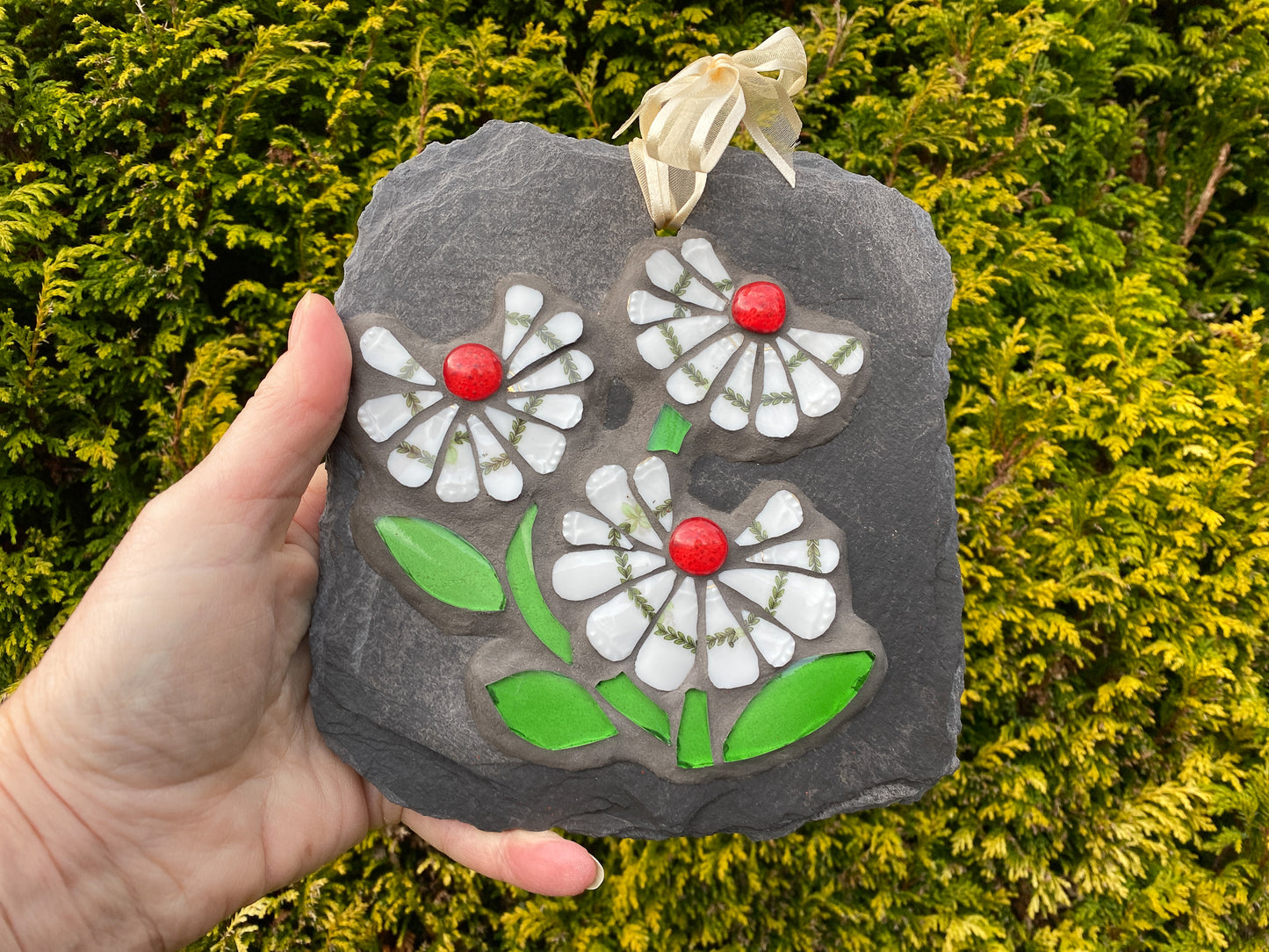 three white daisy flowers, with a green leafy detail, red glass nuggets and bright green leaves in mosaic on slate hand held against an evergreen background
