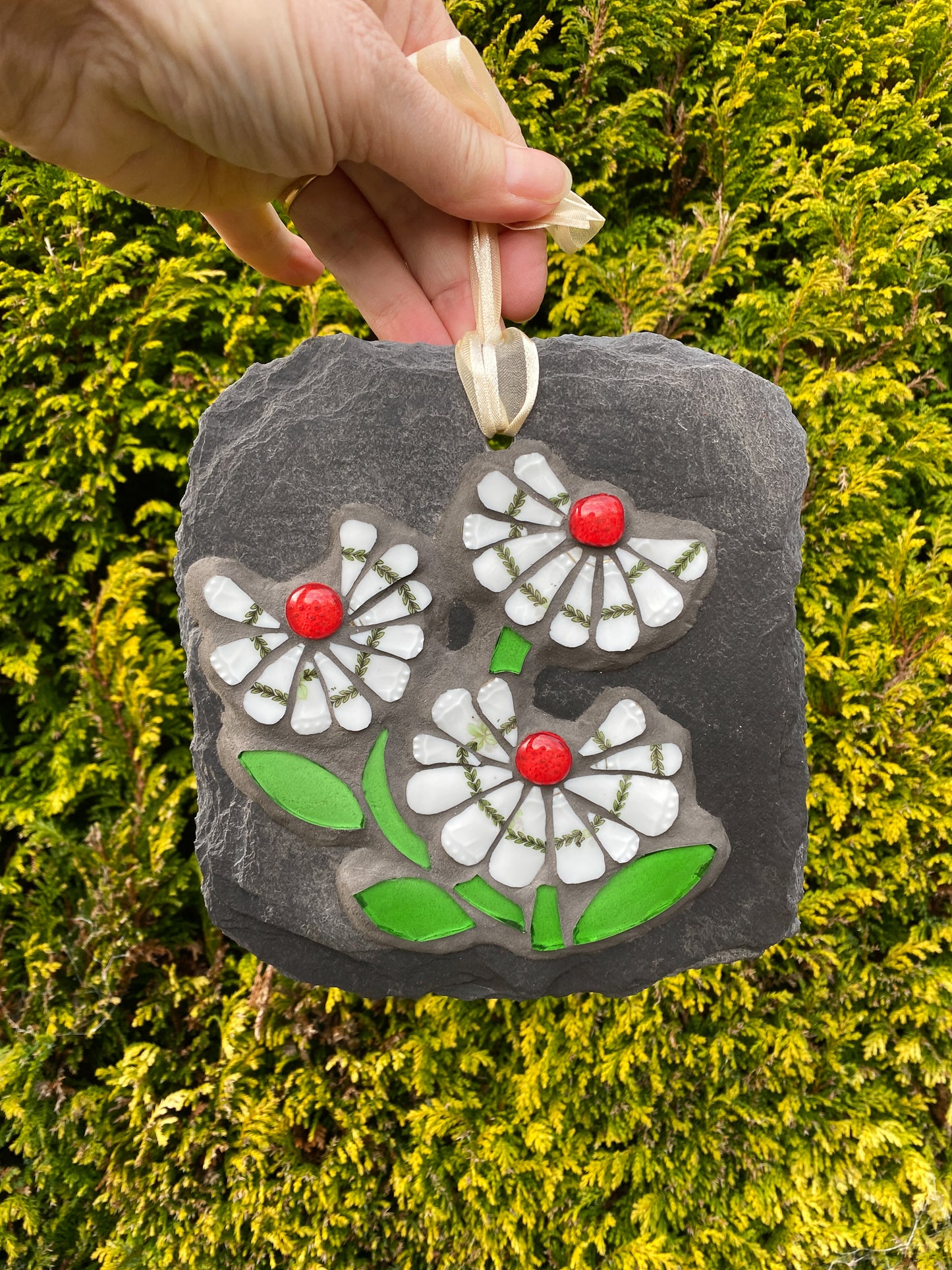 three white daisy flowers, with a green leafy detail, red glass nuggets and bright green leaves in mosaic on slate against an evergreen background