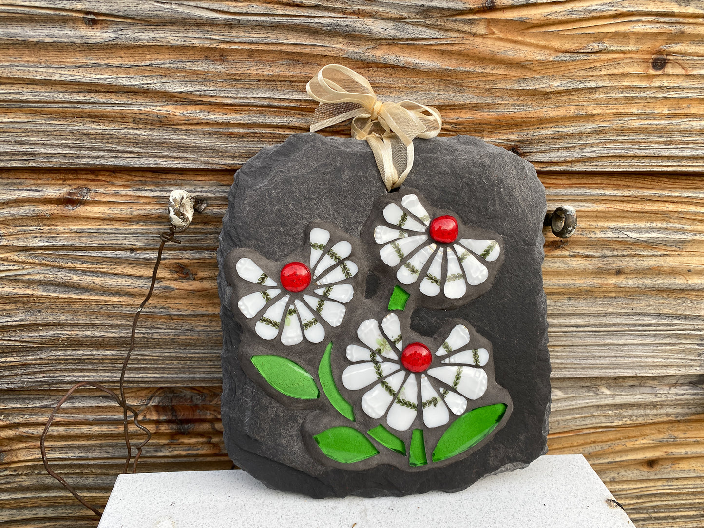 three white daisy flowers, with a green leafy detail, red glass nuggets and bright green leaves in mosaic on slate against a wooden shed background