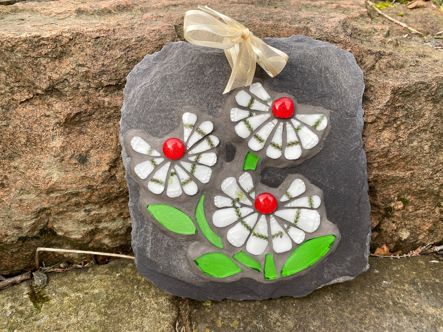 three white daisy flowers, with a green leafy detail, red glass nuggets and bright green leaves in mosaic on slate against a stone background