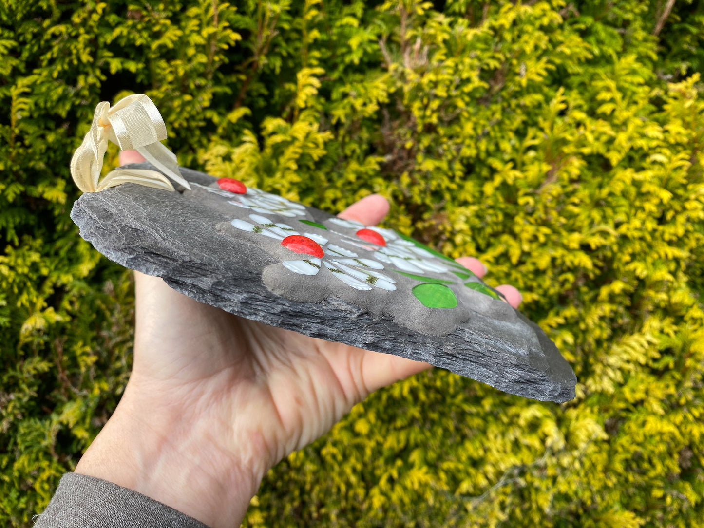 three white flowers and red nuggets in mosaic showing the side of the slate. handheld against an evergreen tree background