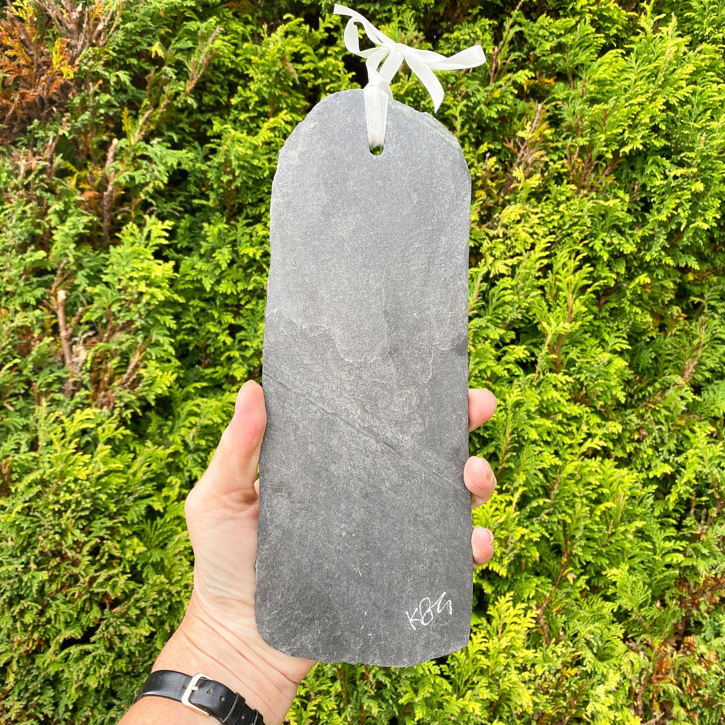 Hand holding a grey slate with a ribbon on it, with 'kbg' signature etched on it against a green bush background