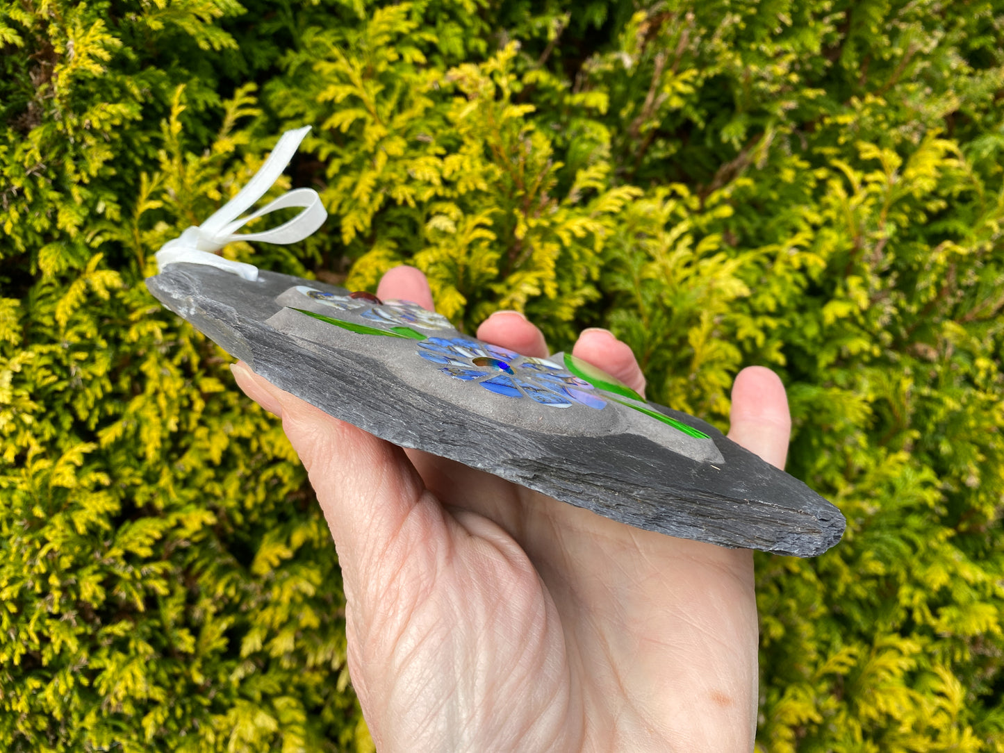 a flower slate hand held showing the side of the slate, against an evergreen tree