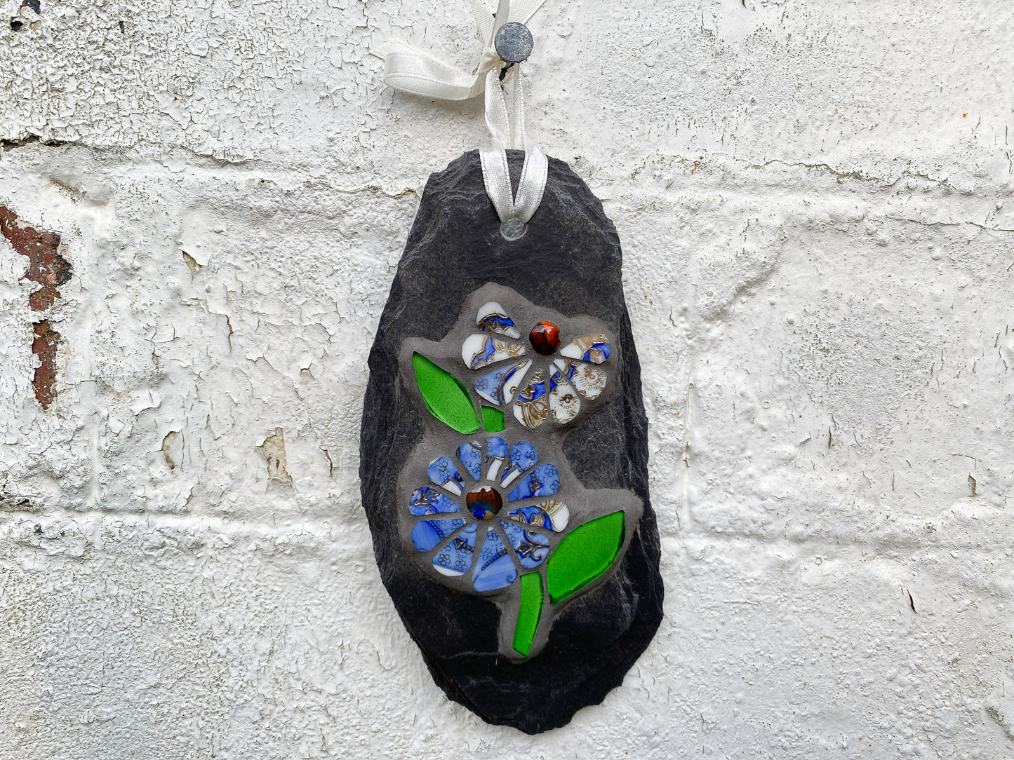 two cornflower blue and white flowers with bright green leaves in mosaic on a grey slate, hand held, with a dustressed brick background