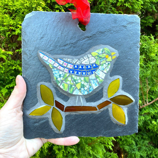Mosaic bird on a branch in blues and greens, on a slate background, shown handheld against an evergreen tree