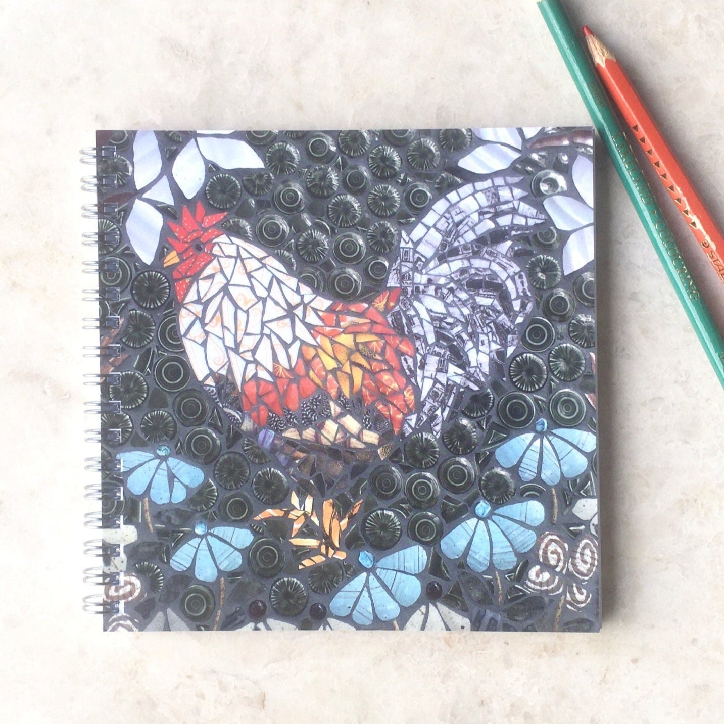 spiral bound notebook and two pencils on a beige background.  the image on the front of the notebook is of a chicken in mosaic