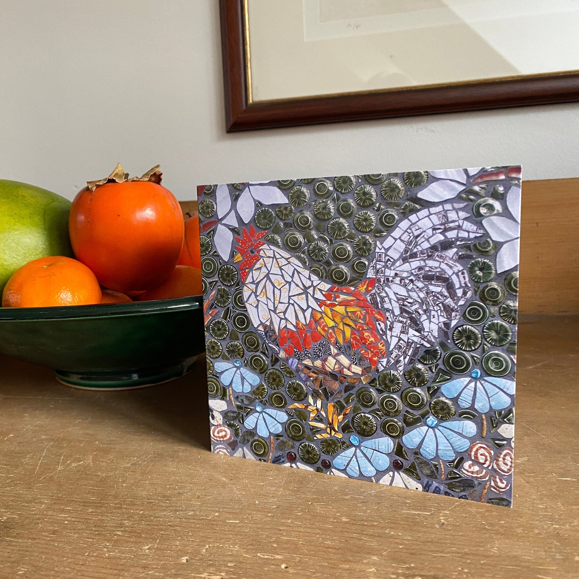 a card with a chicken or cockerel on it, shown beside a bowl of fruit