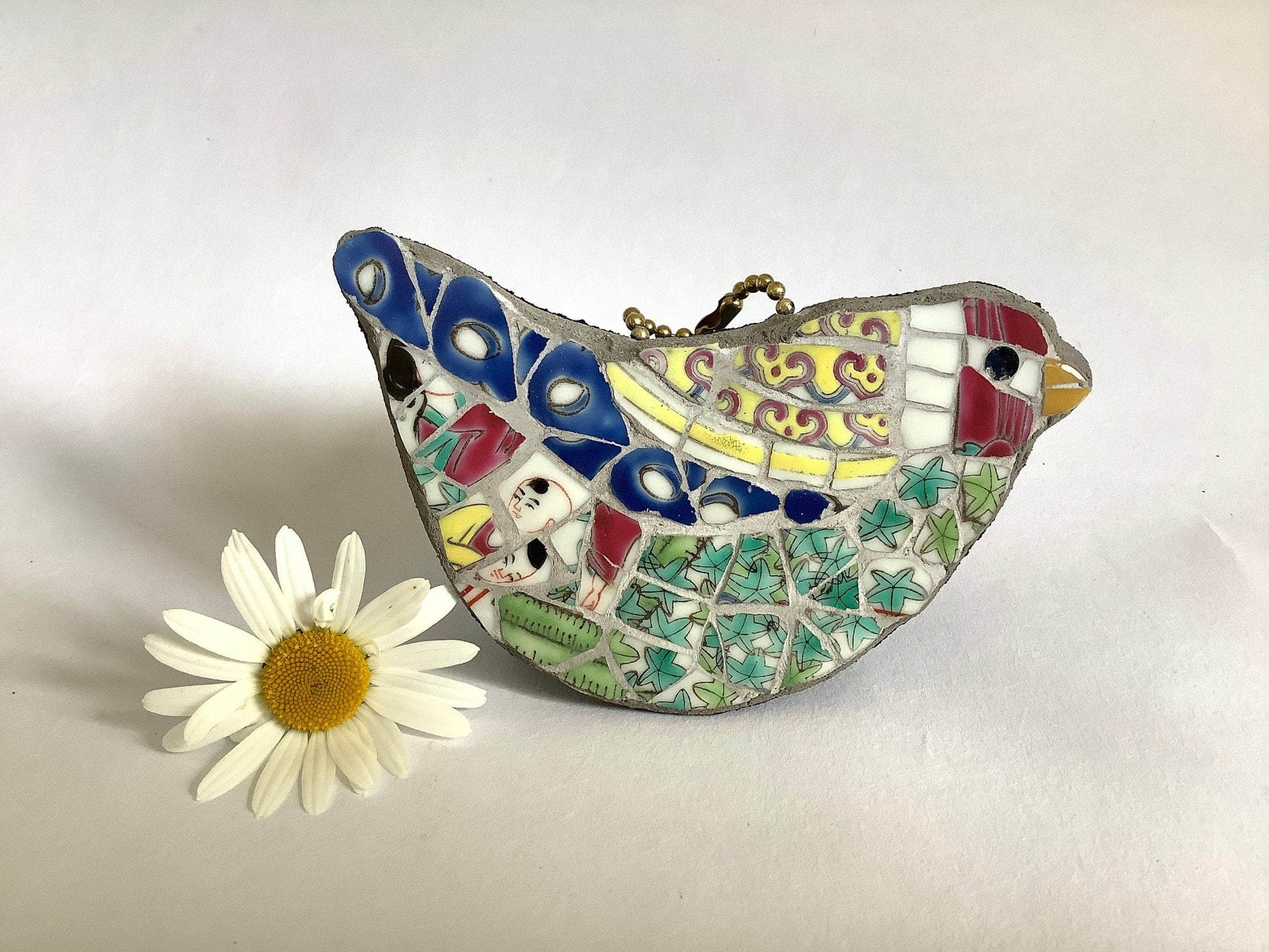 a multicoloured bird mosaic which hangs from a ball chain loop, with a red head, green body and blue feathers, all made from a chinese porcelain pot, including some faces. photographed alongside a white daisy for scale