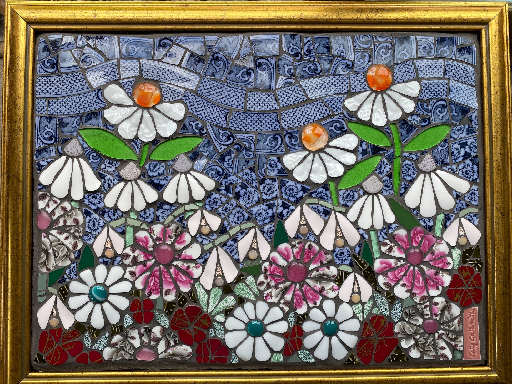 Mosaic artwork of flowers in a gold frame.  White flowers with orange centres, pink and white flowers, blue sky made from crockery