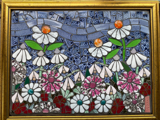 Mosaic artwork of flowers in a gold frame.  White flowers with orange centres, pink and white flowers, blue sky made from crockery