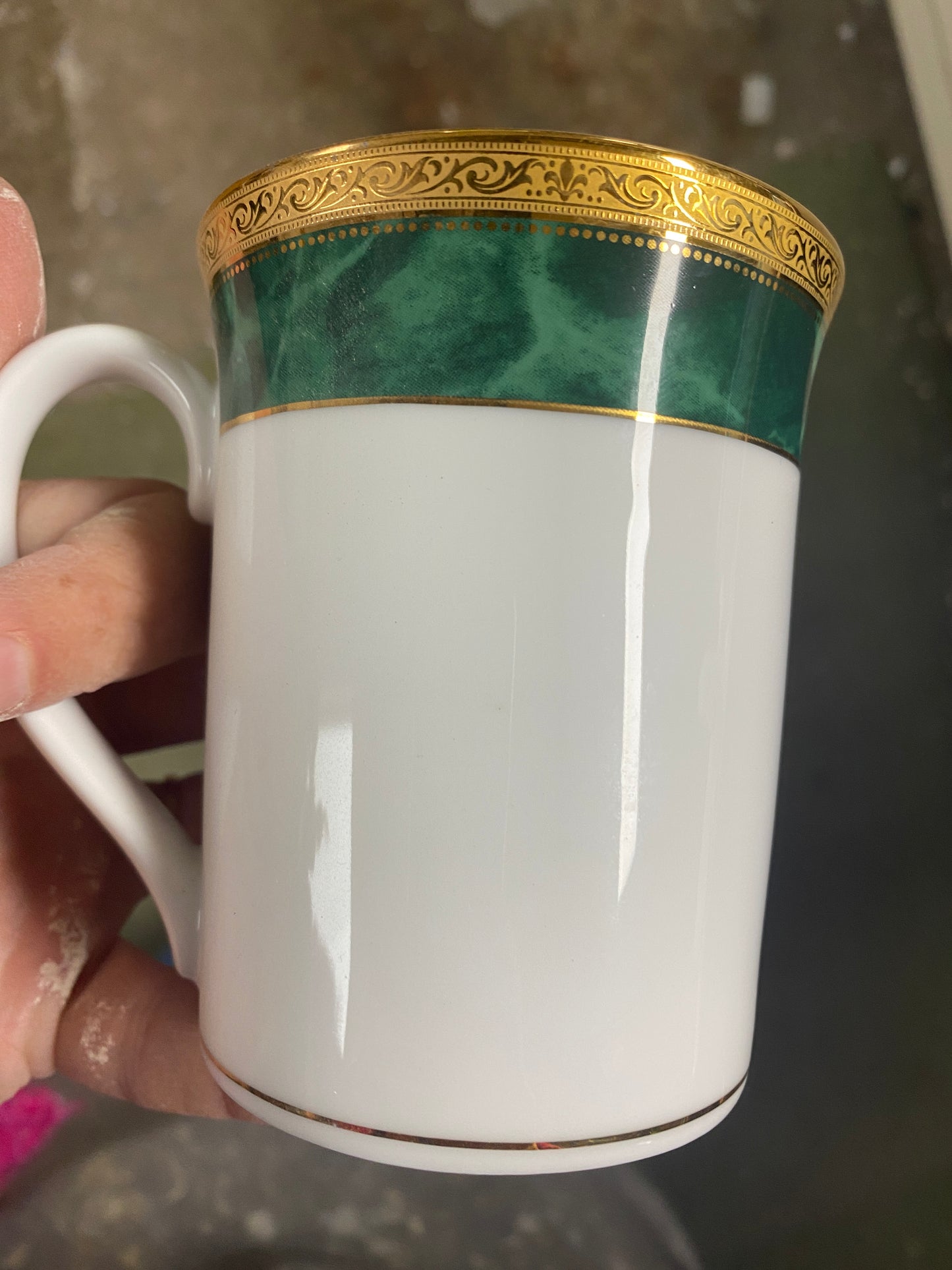 White mug with gold and green decorative band held by a hand against a blurred background