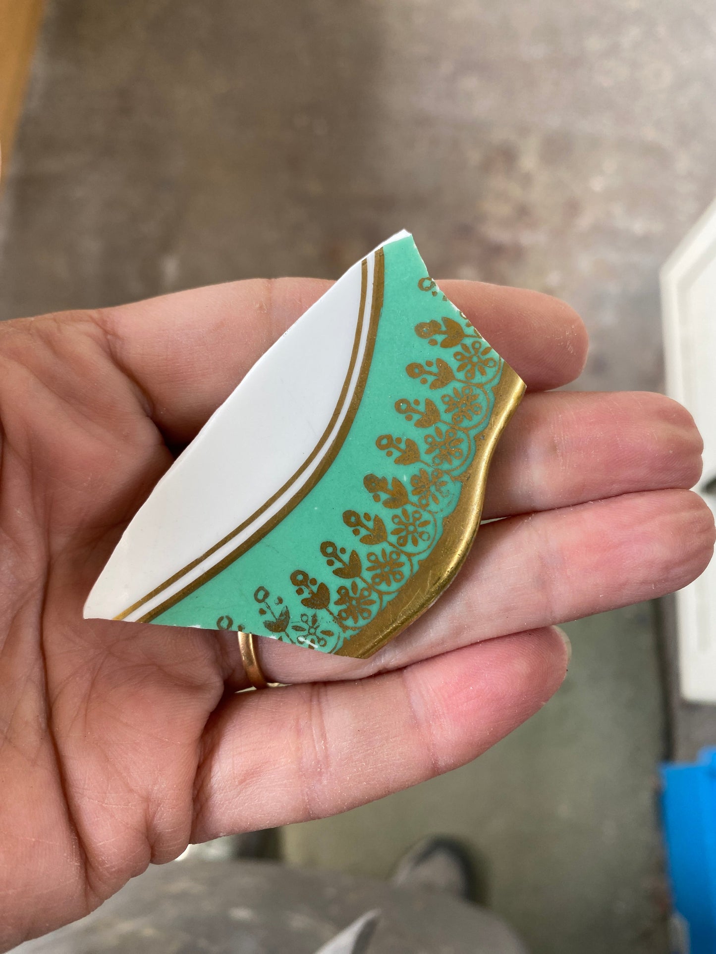 Hand holding a decorative teal and gold ceramic shard against a blurred background