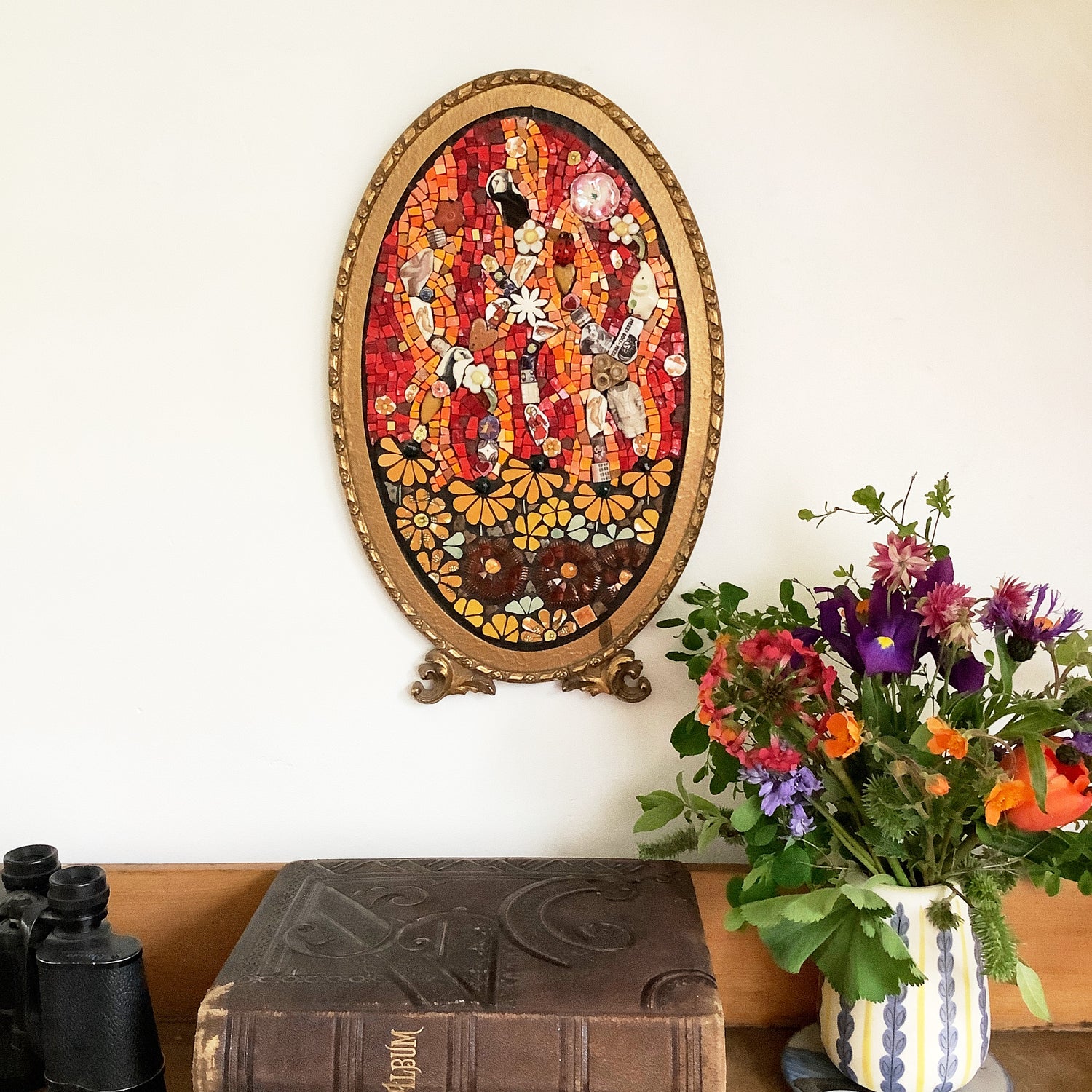 colourful mosaic with flowers in recycled materials at the bottom, and a mosaic in red and orange smalti above.  displayed on a wall with flowers and a book beside