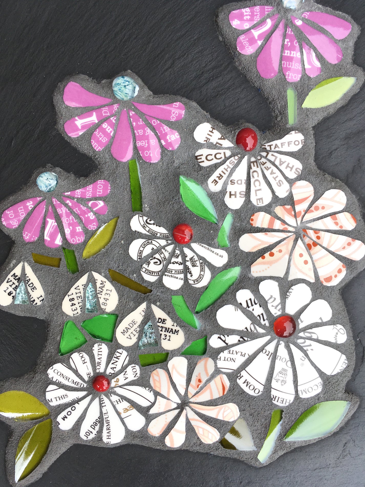 detail of mosaic flowers on slate. the flowers all have text on them, from discarded crockery. Each flower has a glass nugget at the centre of it. 