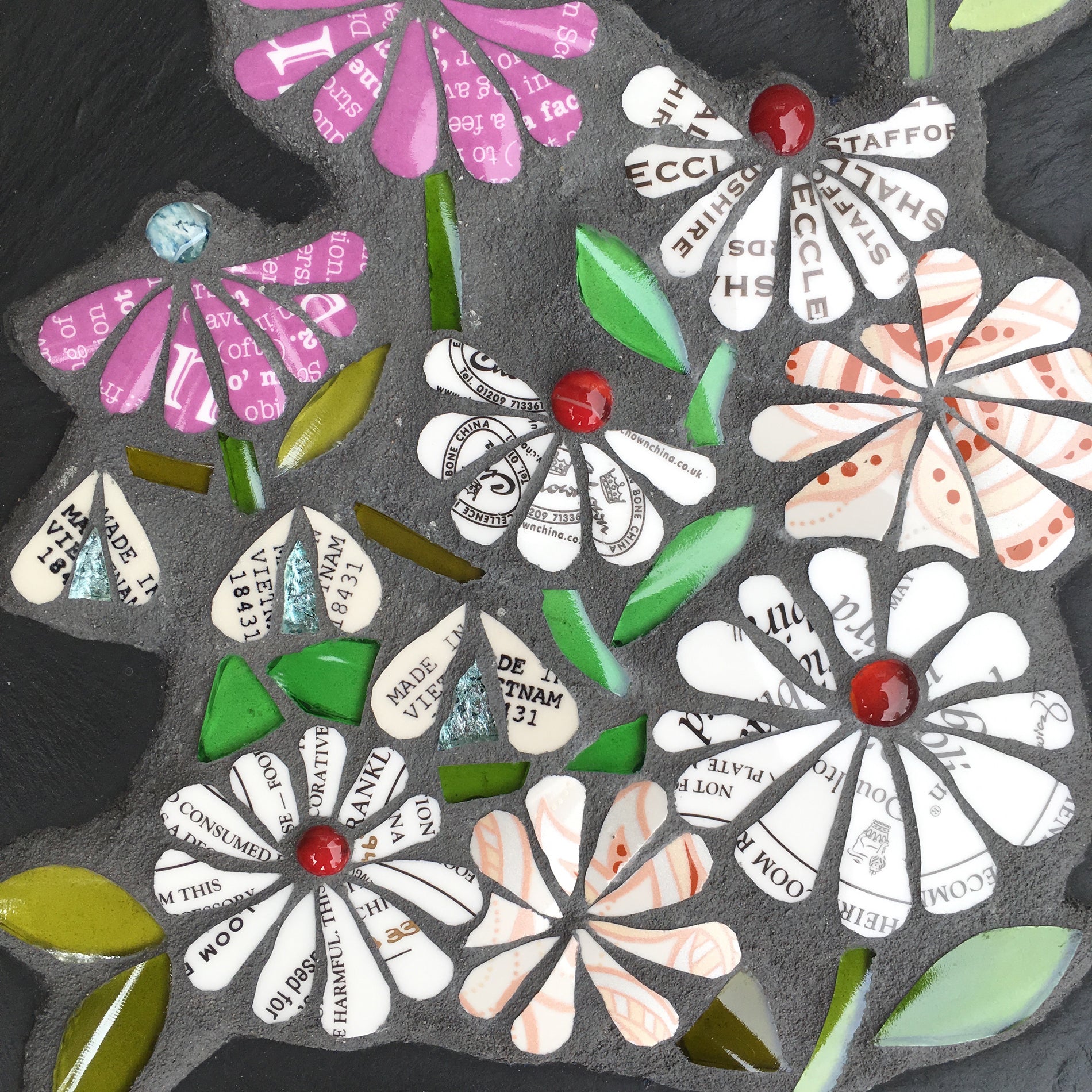 detail of mosaic flowers on slate. the flowers all have text on them, from discarded crockery. Each flower has a glass nugget at the centre of it. 