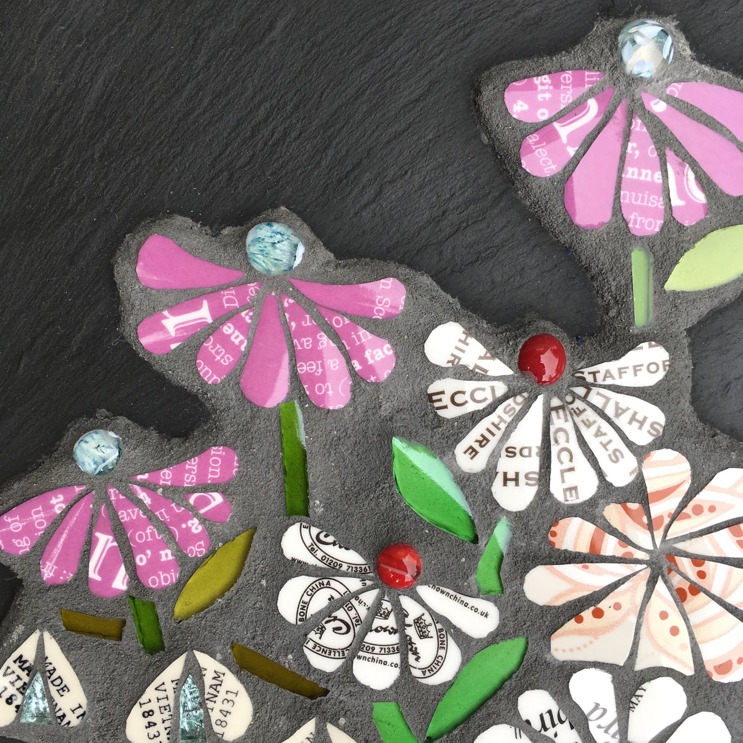 detail of mosaic flowers on a slate. the flowers all have text on them, from discarded crockery. Each flower has a glass nugget at the centre of it. 