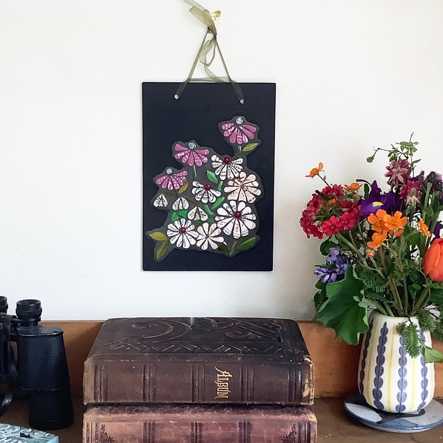 mosaic flowers on a rectangular slate mat. the flowers all have text on them, from discarded crockery. Each flower has a glass nugget at the centre of it. photographed alongside books and flowers