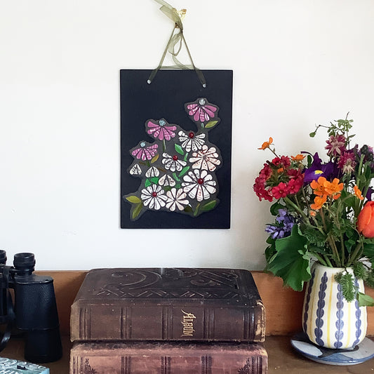 mosaic flowers on a rectangular slate mat. the flowers all have text on them, from discarded crockery. Each flower has a glass nugget at the centre of it. photographed alongside books and flowers
