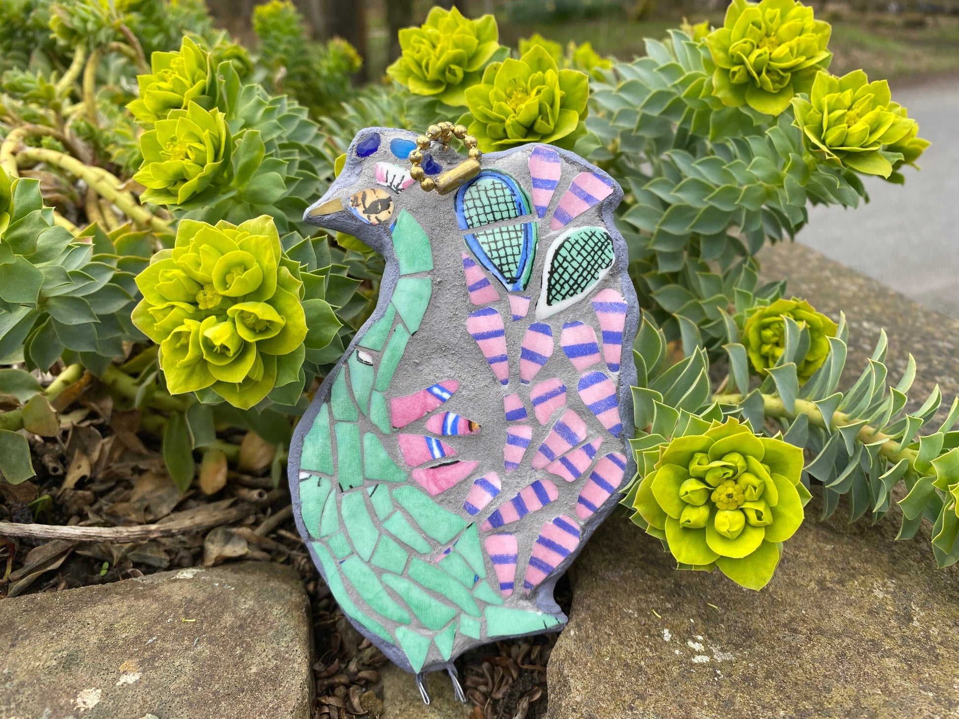 Mosaic peacock as viewed from the side rather than full feather display, with a green body, and pink and blue stripy tail feathers, with a pair of tennis racket heads in the tail.  Made from crockery. photographed with a foliage background