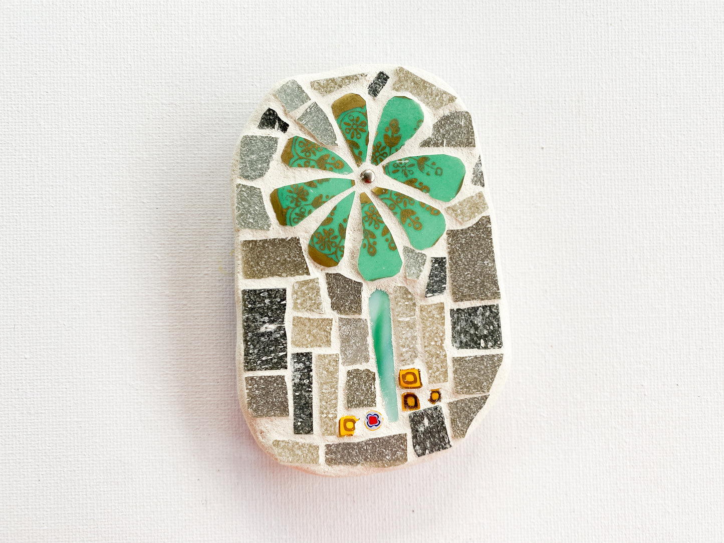 Small mosaic of green and gold petaled flower with grey tile background with millefiori, photographed on white