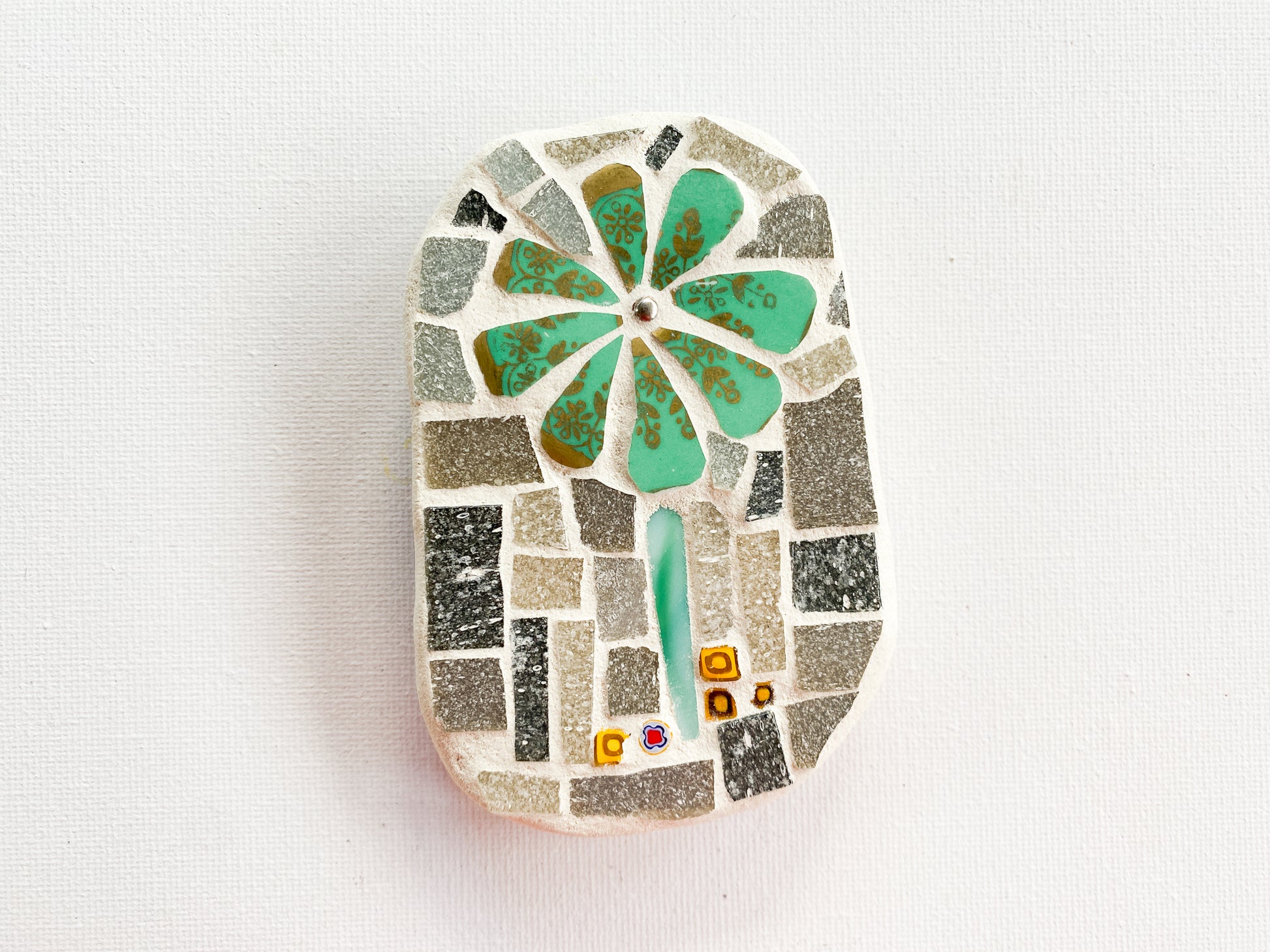 Small mosaic of green and gold petaled flower with grey tile background with millefiori, photographed on white
