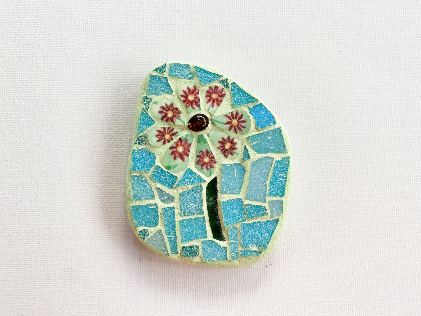Small mosaic of green floral petaled flower with pale blue tile background, photographed on white