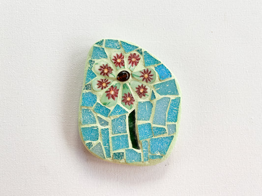 Small mosaic of green floral petaled flower with pale blue tile background, photographed on white