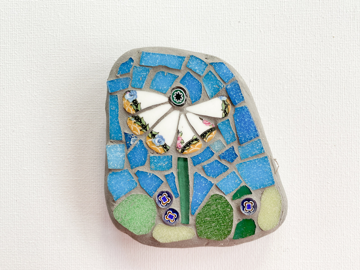 Small mosaic of white petaled flower with blue tile background, with millefiori photographed on white