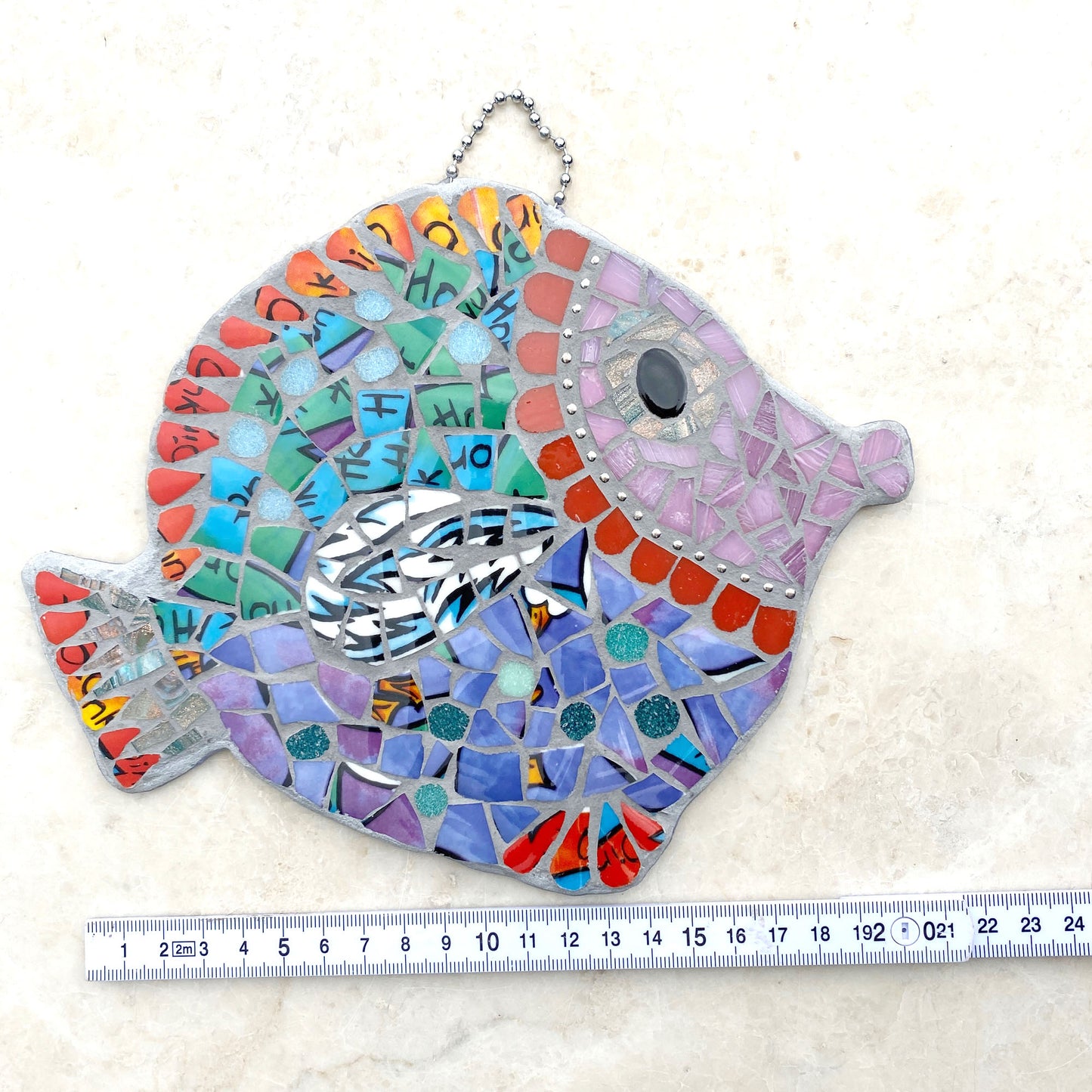 A mosaic fish made from bright purple and turquoise crockery, pink face surrounded by ball chain and curved red tiles.  Fins and tails are in a red china.  Hangs by a ball chain at the top.  Photographed with a ruler, showing length f approx 21cm.