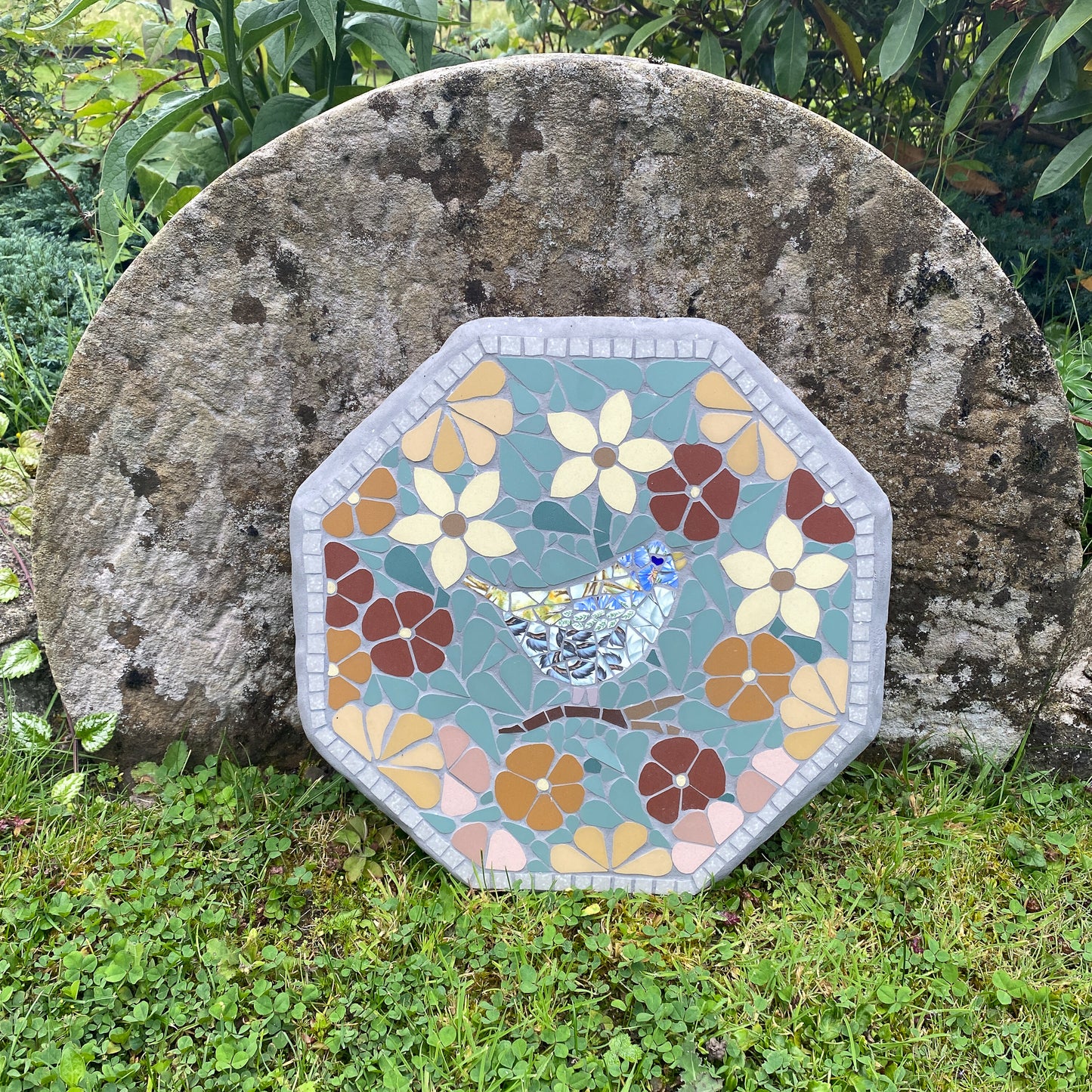 An octagonal mosaic garden stone featuring a bird and flowers, leaning against a large natural rock in a grassy outdoor garden setting.