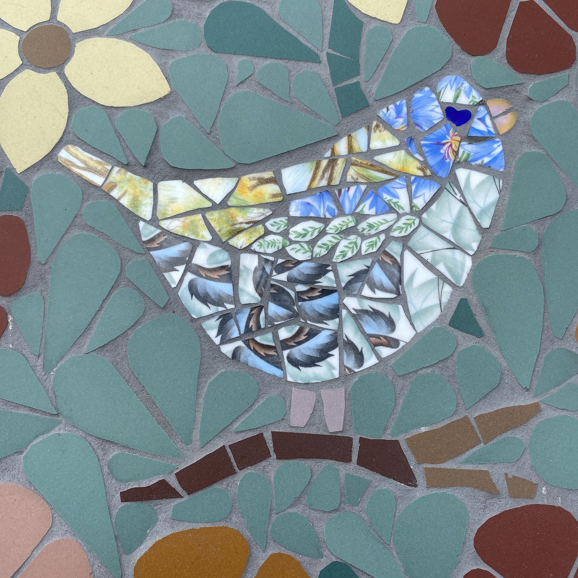 A colorful mosaic bird artwork made from broken ceramic pieces in blue, white, gray, brown, and teal, set against a matching mosaic background with a flower and leaf shapes.
