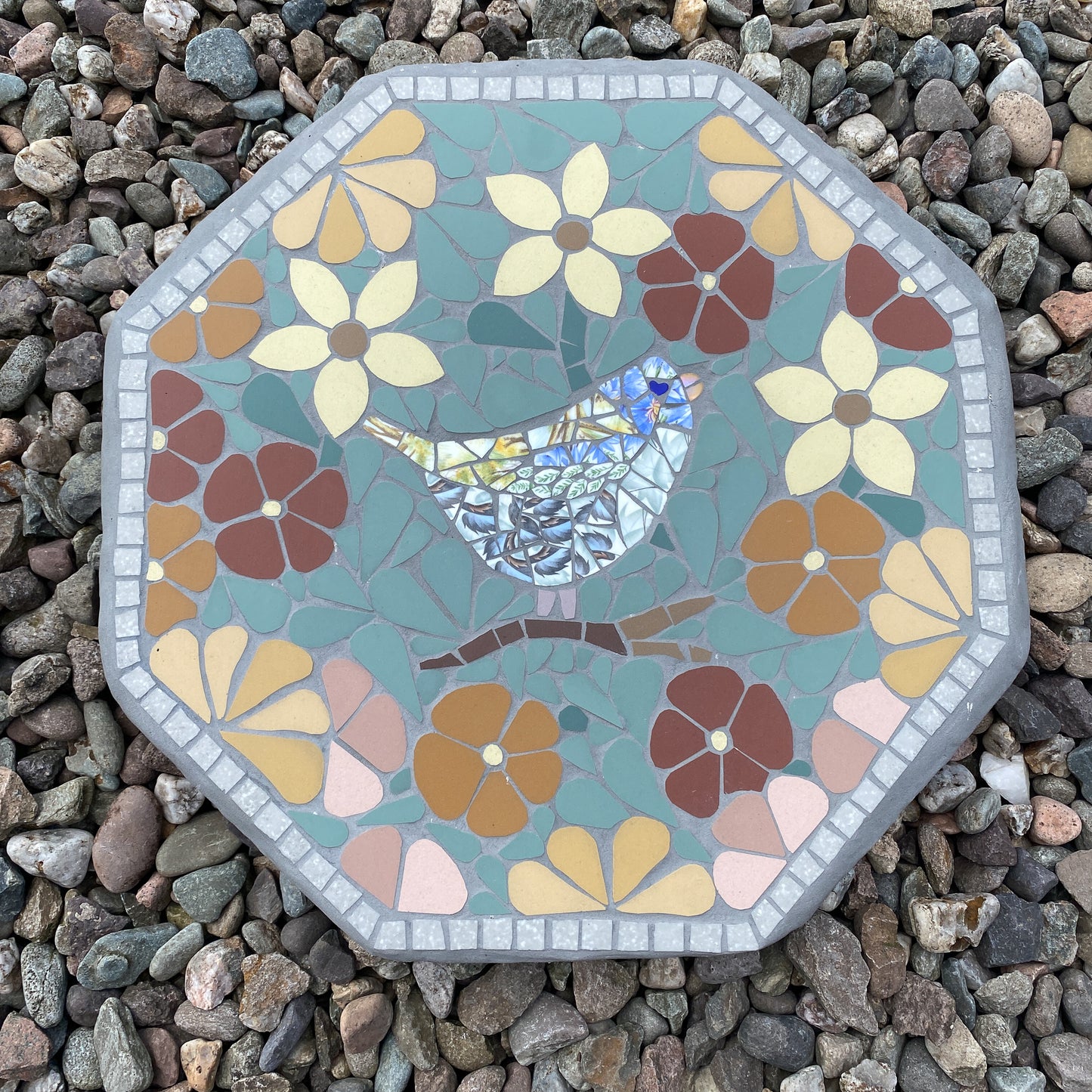 An octagonal mosaic garden stone featuring a bluebird design surrounded by stylized flowers, placed on a bed of small gray pebbles.