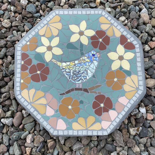 An octagonal mosaic garden stone featuring a bluebird design surrounded by stylized flowers, placed on a bed of small gray pebbles.