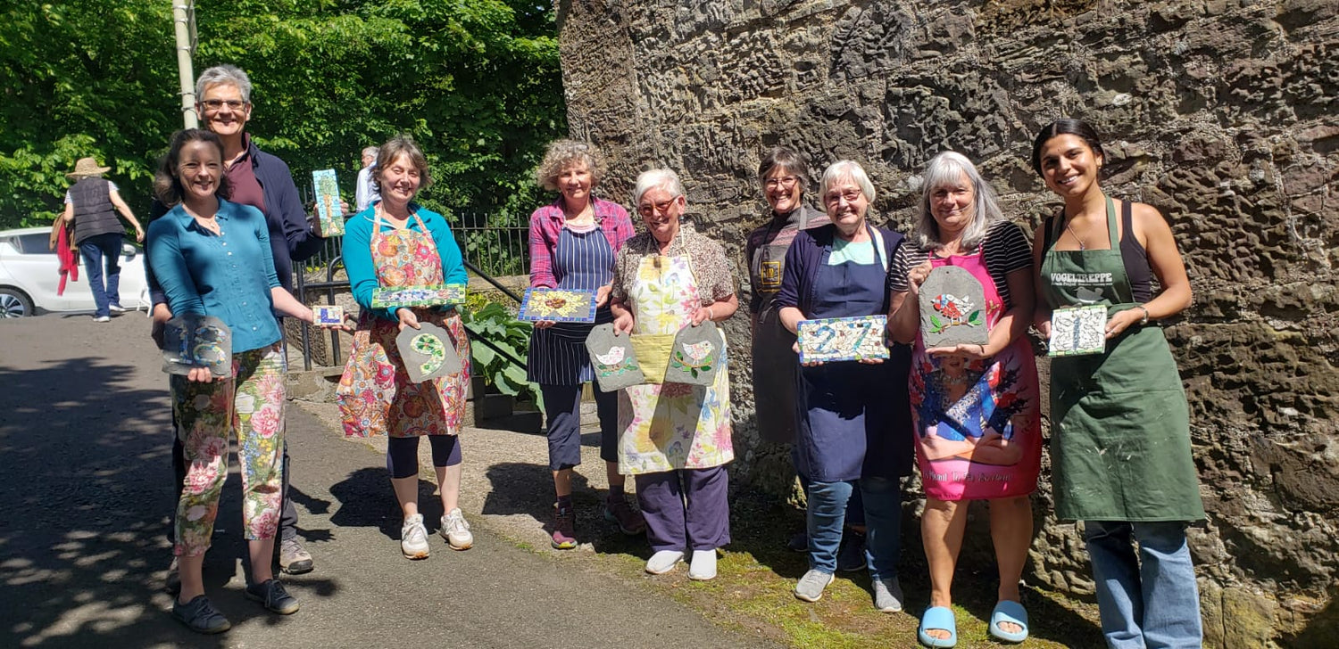 Group of workshop participants holding their completed mosaics in a garden setting.