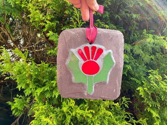 Red and green thistle in mosaic on a grey slate, being held by a ribbon at the top.