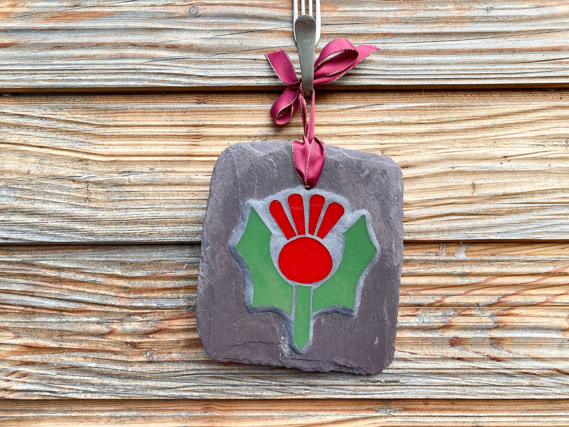 Red and green thistle in mosaic on a grey slate.  Hanging against a horizontal slatted wooden wall
